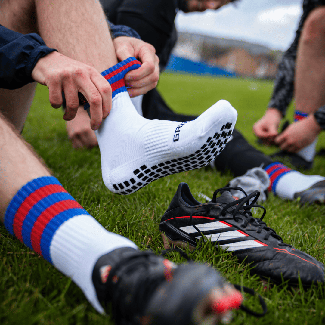 Blue and red GRIPTEC Grip Socks worn with football boots on a pitch