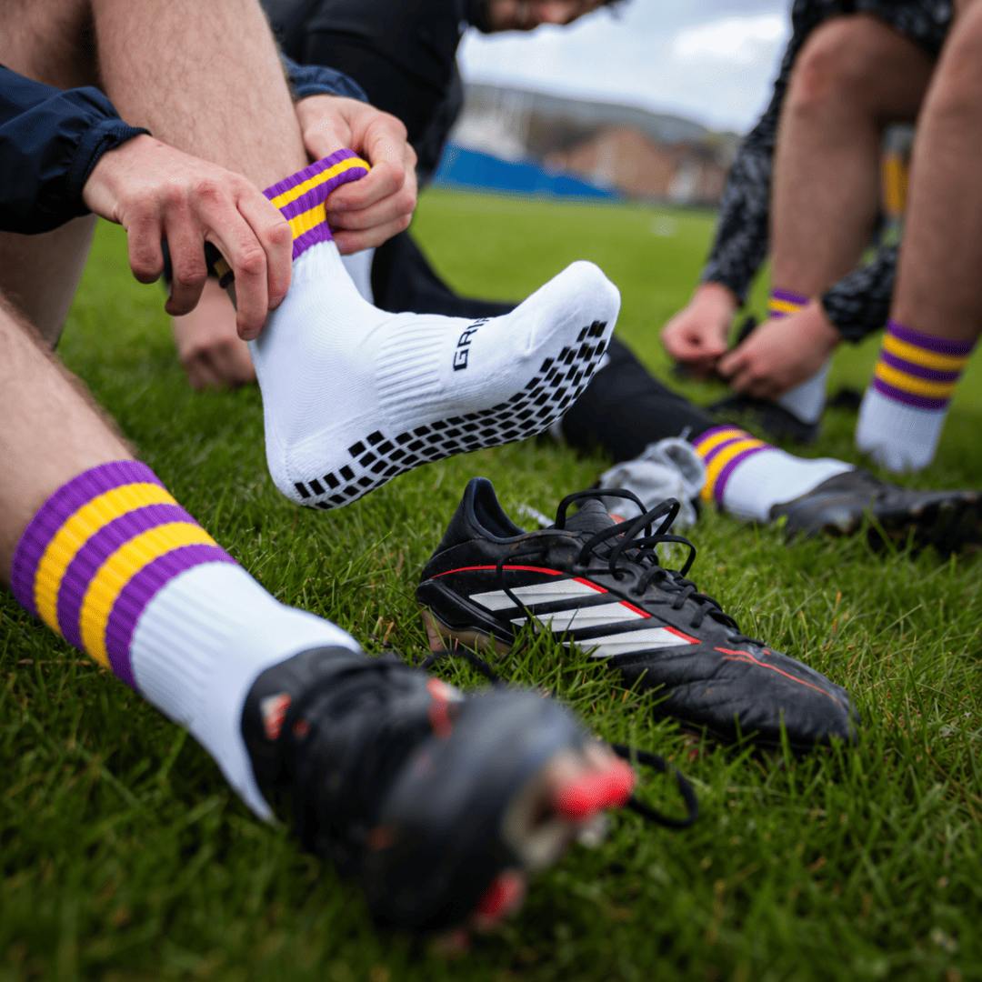 Purple and yellow GRIPTEC Grip Socks worn with football boots on a pitch
